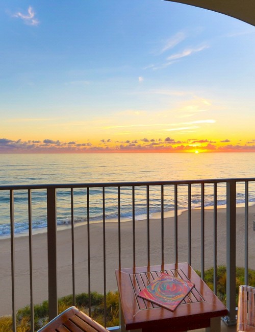 A beachside view at sunset from a balcony with wooden chairs and a small table, overlooking the ocean and sandy shore.