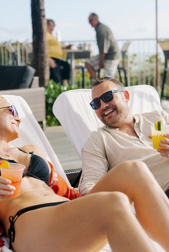 Two people are lounging on chairs by a pool, enjoying drinks and smiling at each other with more people in the background.