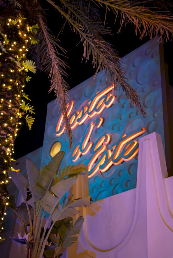 A neon sign is displayed against a textured blue wall with tropical plants and palm branches lit by decorative lights.