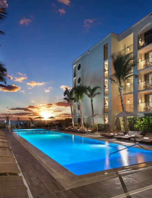 A resort pool area at sunset with lounge chairs, umbrellas, palm trees, and a multi-story building with balconies in the background.