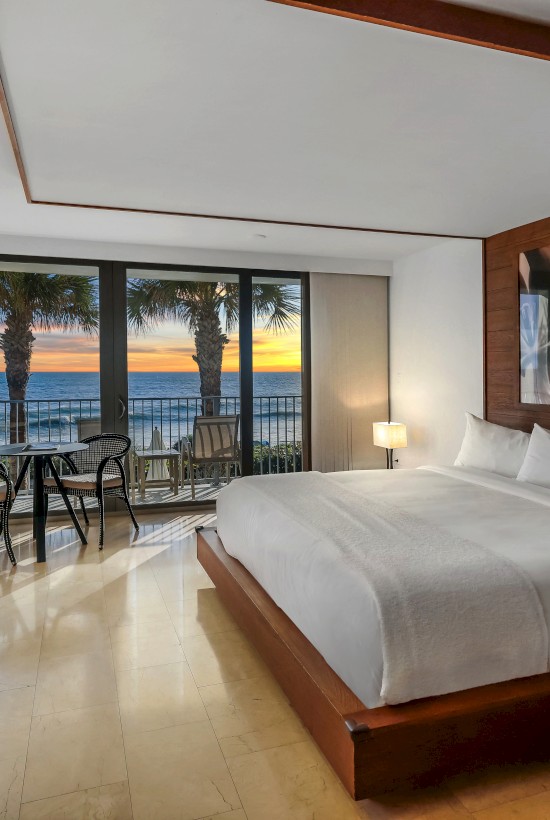A modern hotel room with a large bed, TV, balcony view of the ocean and sunset, palm trees, and cozy seating area.