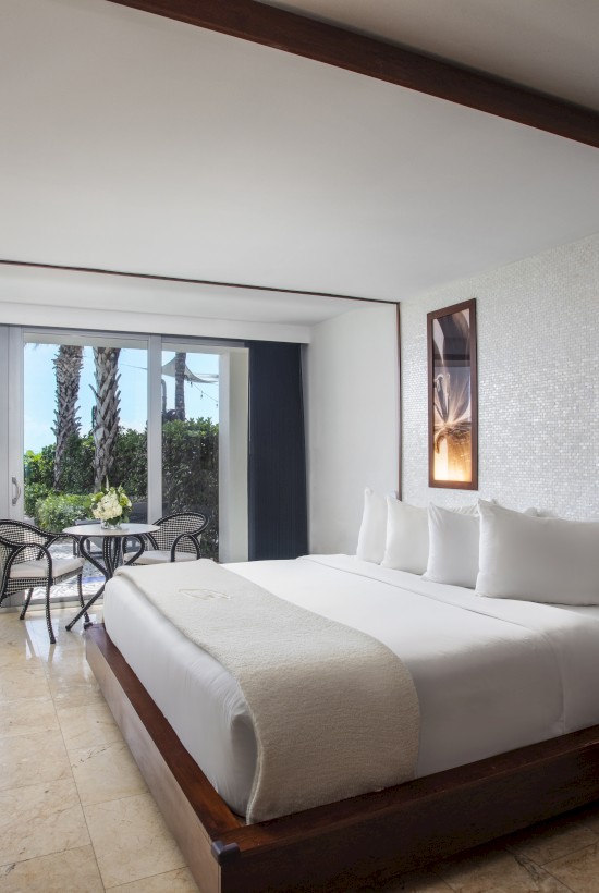 A modern hotel room with a large bed, TV, and seating area, opening to a patio with greenery and partial ocean view.
