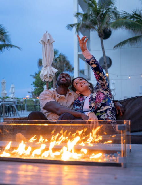 A couple sitting by a fire pit, smiling and taking a selfie in an outdoor setting with palm trees in the background.