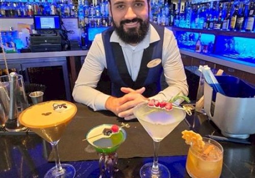 A bartender stands behind a bar with four colorful cocktails in front of him, in a dimly lit setting.