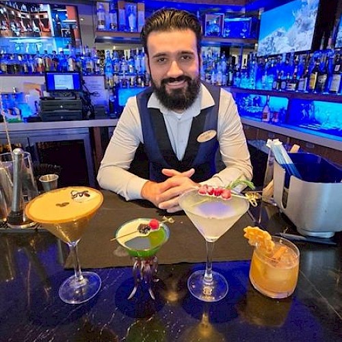 A bartender stands behind a bar with four colorful cocktails in front of him, in a dimly lit setting.