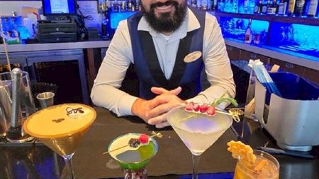A bartender stands behind a bar with four colorful cocktails in front of him, in a dimly lit setting.