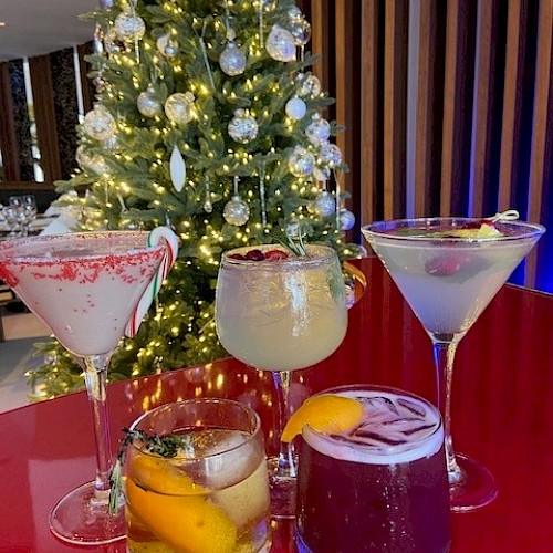 A variety of holiday cocktails placed on a red piano top, with a decorated Christmas tree in the background.