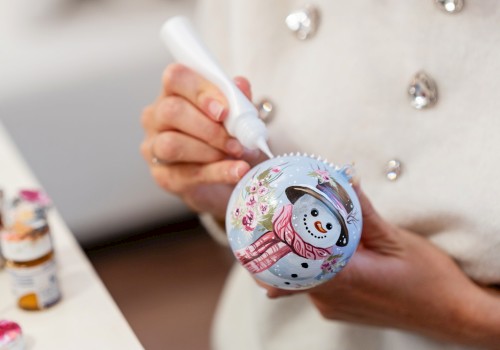 A person decorates a Christmas ornament with a snowman design using a white paint marker, with paint bottles visible in the background.