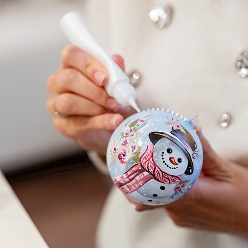 A person decorates a Christmas ornament with a snowman design using a white paint marker, with paint bottles visible in the background.