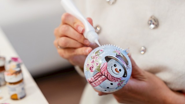 A person decorates a Christmas ornament with a snowman design using a white paint marker, with paint bottles visible in the background.