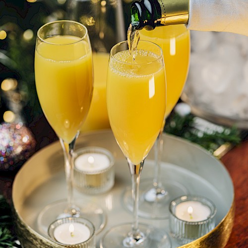Glasses of orange juice mimosas are being poured from a champagne bottle on a tray with candles, set against a festive holiday backdrop.