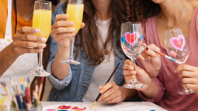 Three friends celebrate with painted glass crafts and bright drinks, sharing smiles and creative arts around a colorful table.