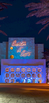 A night scene at a resort with palm trees lit by string lights and a blue-lit entrance, Costa del Este sign, and decorative arches.