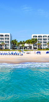 Two white beachfront buildings with palm trees, beach chairs, and umbrellas line a sandy beach meeting clear turquoise ocean waters.