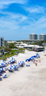 The image shows a sunny beach with blue umbrellas, people relaxing, and high-rise hotels along the shoreline under a partly cloudy sky.