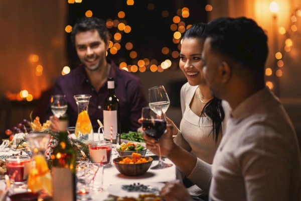 A group of friends toasting at a warm dinner party with wine, candles, and festive lights in the background.