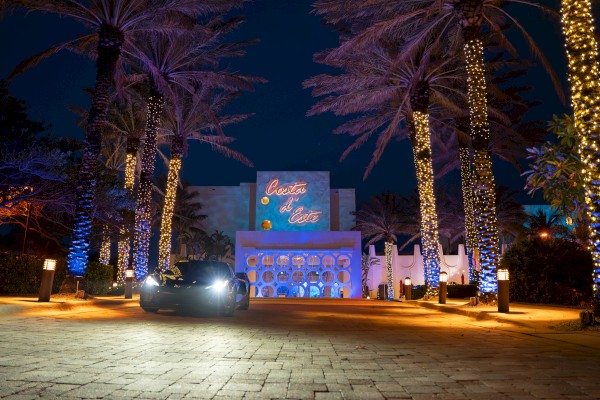 A nighttime hotel entrance with palm trees wrapped in warm lights, a blue-lit facade, and a car headlights shining toward the front.