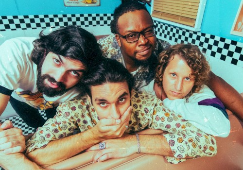 Four friends crowded on a pink couch in a retro diner, posing playfully for a photo with bright blue walls and checkered accents.