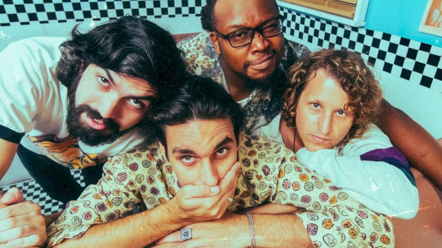 Four friends crowded on a pink couch in a retro diner, posing playfully for a photo with bright blue walls and checkered accents.