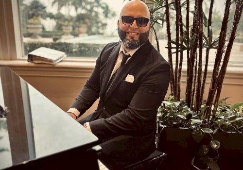 A person in a suit and sunglasses plays a grand piano indoors, with tall plants and a large window showing an outdoor scene in the background.