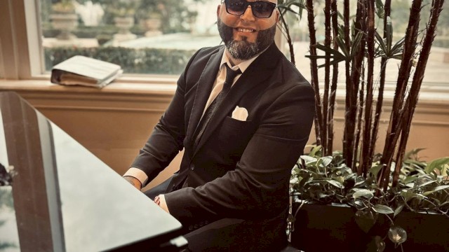 A person in a suit and sunglasses plays a grand piano indoors, with tall plants and a large window showing an outdoor scene in the background.