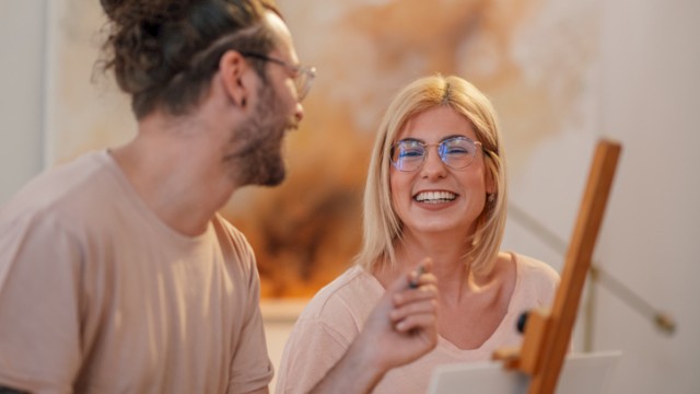 Two friends painting together, smiling and chatting in a cozy studio, sharing a moment of creativity and laughter as they work on a canvas.