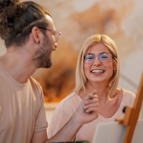 Two friends painting together, smiling and chatting in a cozy studio, sharing a moment of creativity and laughter as they work on a canvas.