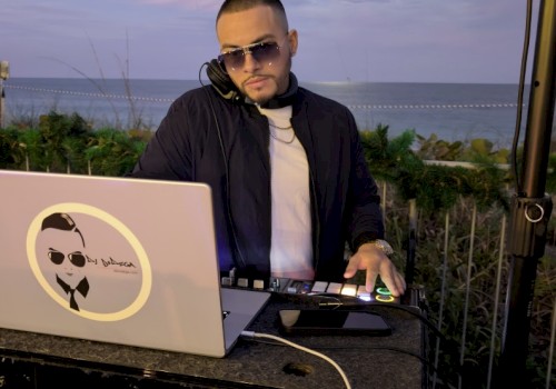 A DJ wearing sunglasses and a black jacket, mixing music on a laptop and turntables by the beach at sunset. End with a period.