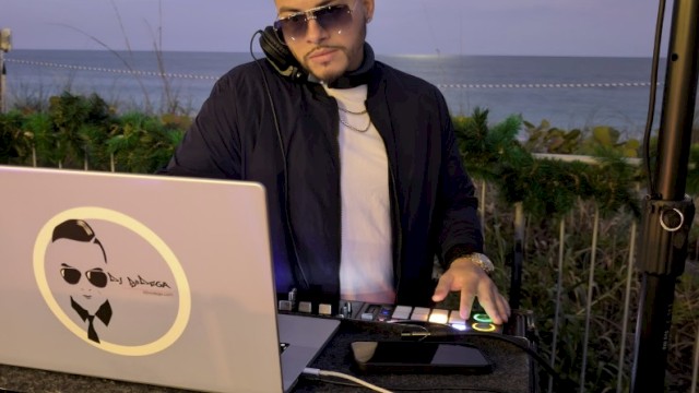 A DJ wearing sunglasses and a black jacket, mixing music on a laptop and turntables by the beach at sunset. End with a period.