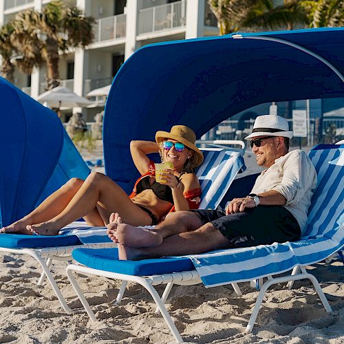 Two people are relaxing on blue lounge chairs under canopies on a sandy beach, with a building and palm trees in the background.