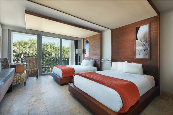 A hotel room with two beds featuring orange throws, a seating area, large windows, and modern decor, with artwork above the beds showing a flower.