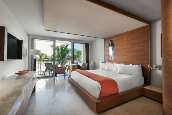 A modern hotel room with a large bed, orange throw, TV, seating area, and a balcony featuring a view of palm trees and the outdoors.