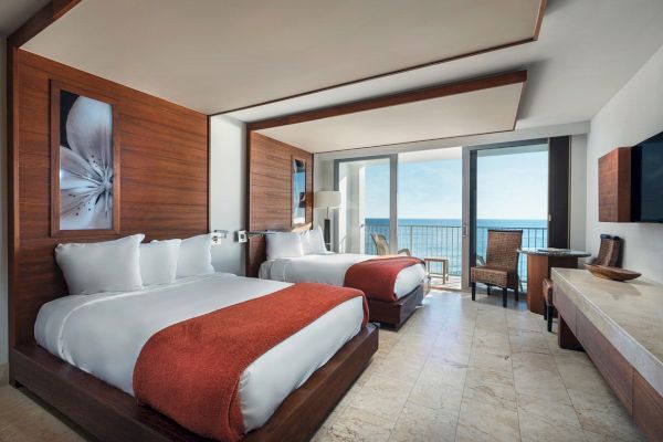 A modern hotel room with two beds, orange throws, wooden furniture, a TV, and a balcony with an ocean view in the background.