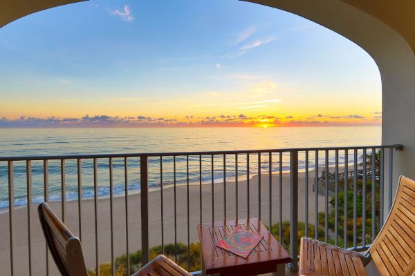 A serene beach view from a balcony at sunset, featuring a table with magazines and two wooden chairs, overlooking the ocean and sandy shore.