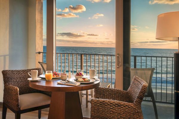 A cozy balcony setup with a round table, wicker chairs, breakfast items, overlooking the ocean at sunset under a partly cloudy sky.