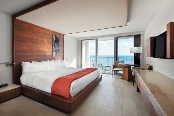 A modern hotel room with a large bed, wooden accents, a desk, a chair, and a view of the ocean through a glass door.