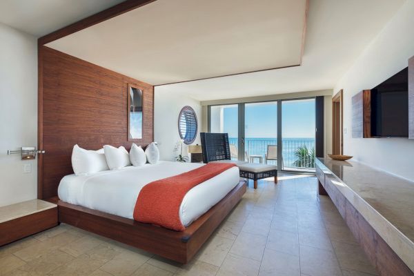A modern hotel room features a large bed with white linens, a red throw, a seating area, and a balcony overlooking the ocean under bright natural light.