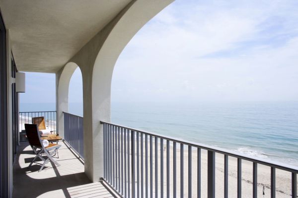 A beachfront balcony with two lounge chairs, a small table, and a stunning view of the ocean under a clear sky, perfect for relaxing.
