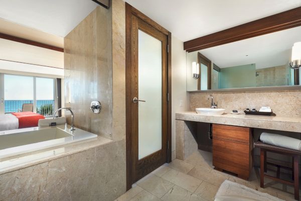 A modern bathroom features a large mirror, wooden accents, a bathtub, sink, and a glimpse of an adjoining bedroom with an ocean view.