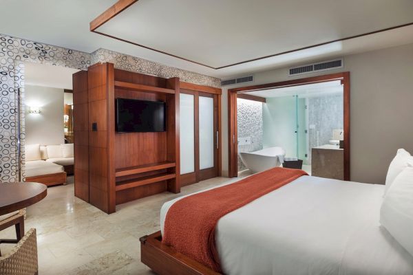 A modern hotel room featuring a large bed, wall-mounted TV, wooden furniture, and a bathroom with a freestanding bathtub separated by glass doors.