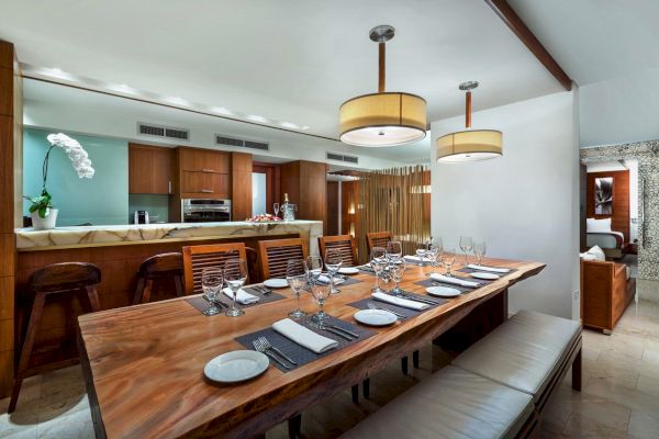 A modern dining room with a large wooden table, six place settings, bench seating, and a kitchen area in the background, decorated with orchids.