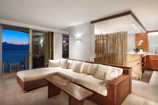 A modern living area features a large sectional sofa, a wooden coffee table, and a kitchenette. There is a balcony offering an ocean view at sunset.