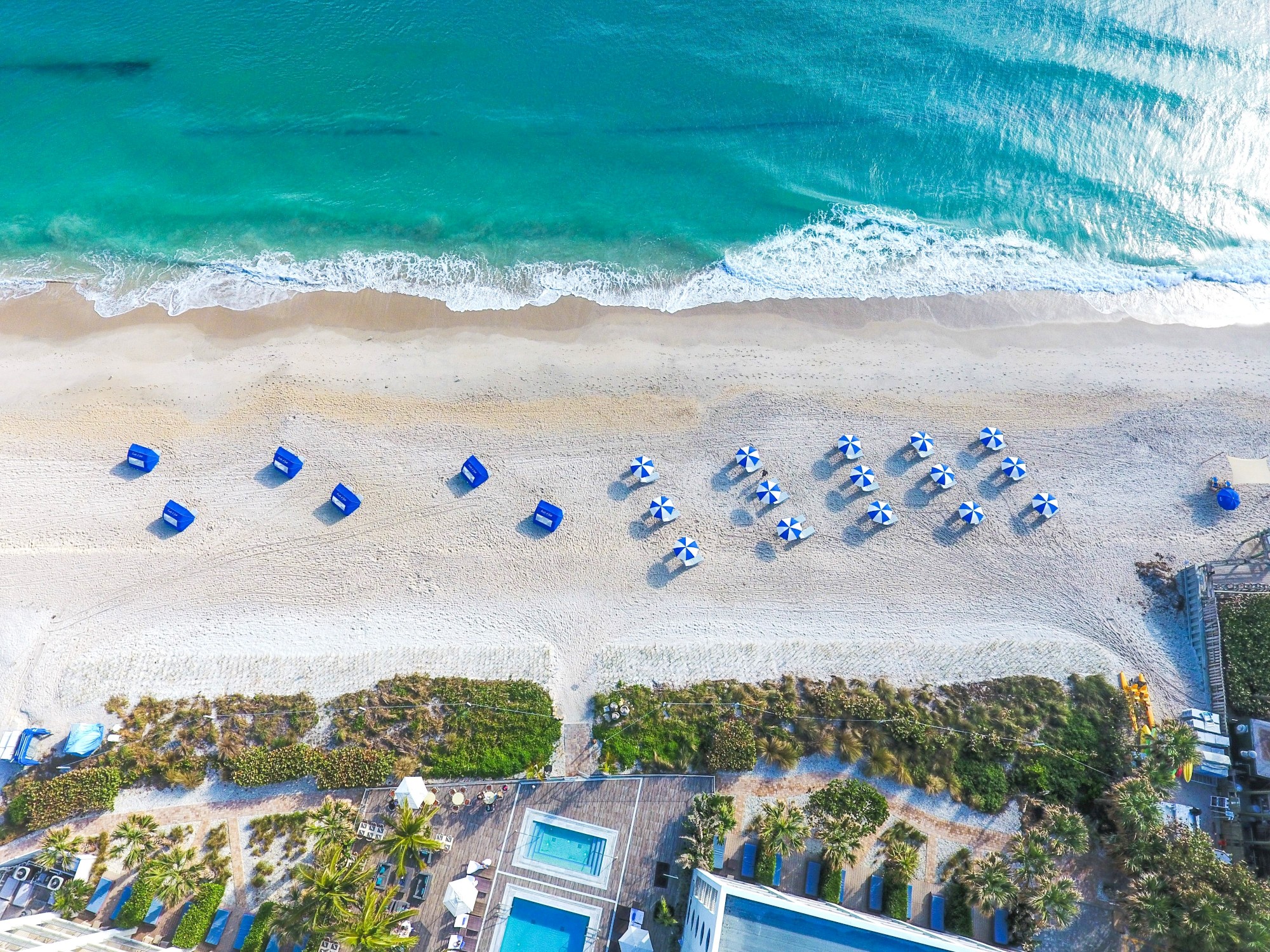 An aerial view of a beach with turquoise water, waves, rows of blue umbrellas, and beach chairs on the sand, with buildings and pools nearby.