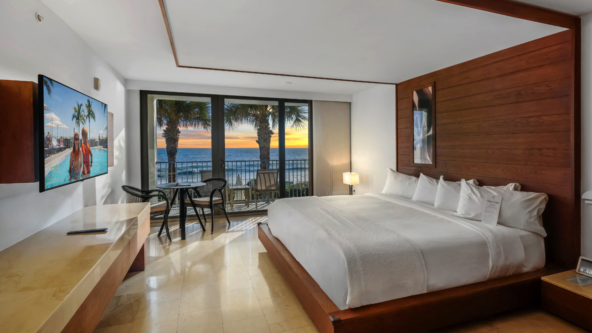 A hotel room with a king-sized bed, TV, and a view of the ocean through glass doors. A small table and chairs are near the window.