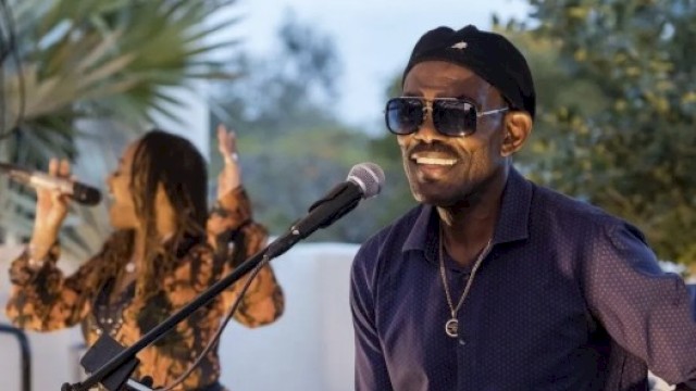 A man wearing sunglasses and a cap plays a shiny steel percussion drum on a sunny outdoor stage, with a female singer in the background.