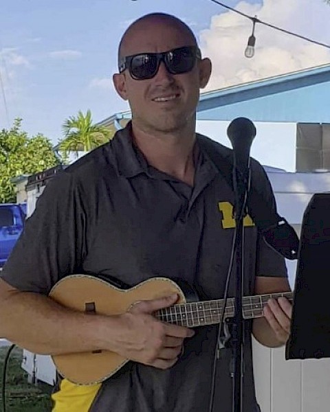 A man with sunglasses is holding a ukulele, standing in front of a microphone, outdoors near a blue building.