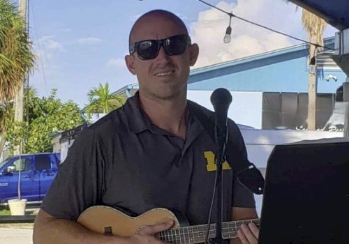 A man with sunglasses is holding a ukulele, standing in front of a microphone, outdoors near a blue building.