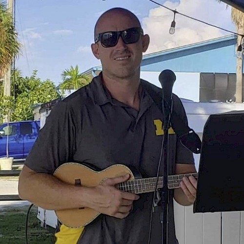 A man with sunglasses is holding a ukulele, standing in front of a microphone, outdoors near a blue building.