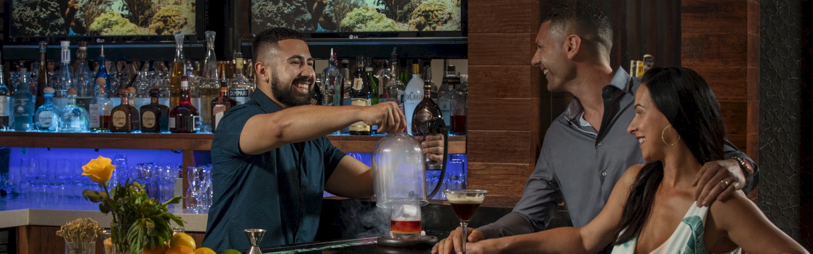 Three people at a bar: a bartender preparing a drink and a couple conversing. There are bottles and screens displaying underwater scenes.