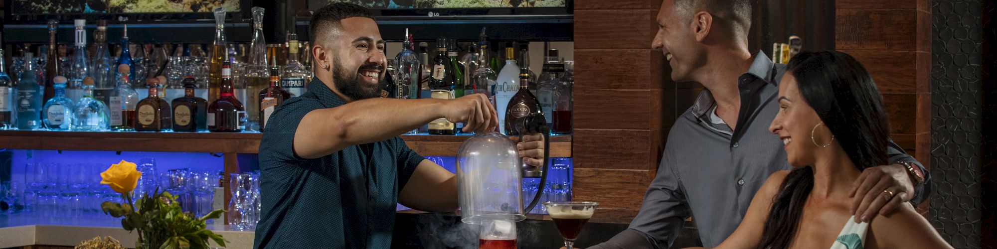 Three people at a bar: a bartender preparing a drink and a couple conversing. There are bottles and screens displaying underwater scenes.